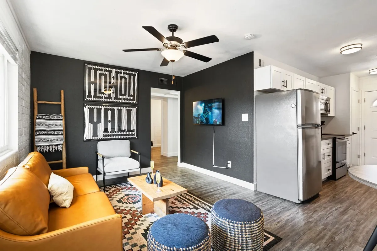 A cozy, contemporary living space featuring a leather sofa, geometric rug, decorative wall art, a wall-mounted TV, and a kitchen area in the background. The room is designed for group gatherings, watching TV, playing games, and sharing meals in a casual, inviting atmosphere.