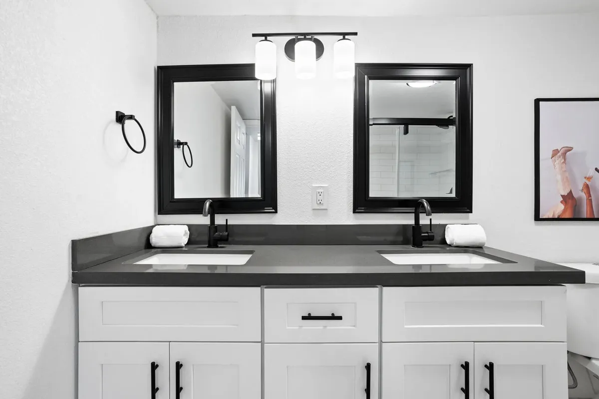 A modern and inviting bathroom featuring double sinks, black-framed mirrors, stylish lighting fixtures, and decorative artwork. The clean lines and ample counter space suggest a well-equipped space for groups to get ready comfortably.