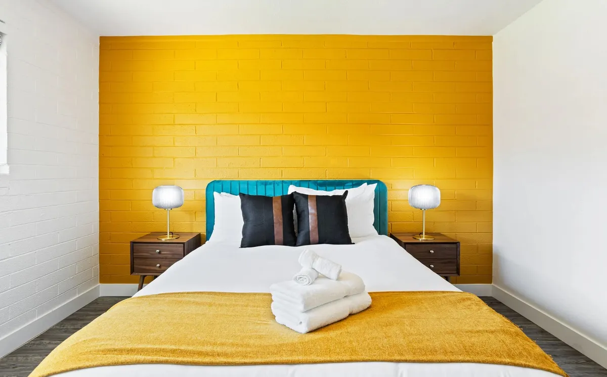 A bright and cheerful bedroom featuring a bold yellow accent wall, a plush bed with a teal headboard and white bedding, wooden nightstands, and table lamps. Warm natural light fills the space, creating a welcoming atmosphere ideal for relaxation and restful nights.