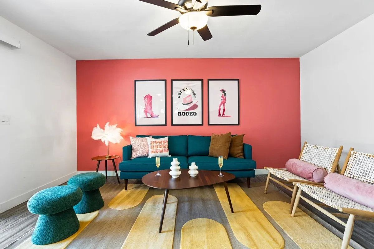 The image shows a bright, cheerful living space featuring a plush teal sofa, colorful artwork on the walls, a modern round coffee table, and unique seating options, creating an inviting atmosphere for socializing and celebrating with friends and family.