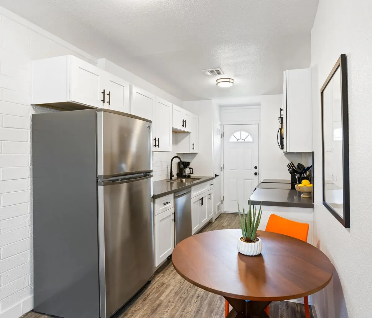 This interior image captures a modern kitchen scene with a stainless steel refrigerator, a round wooden dining table, a bright orange chair, and a potted plant. The kitchen appears well-equipped and ready for cooking and dining activities, with inviting lighting and cozy touches.