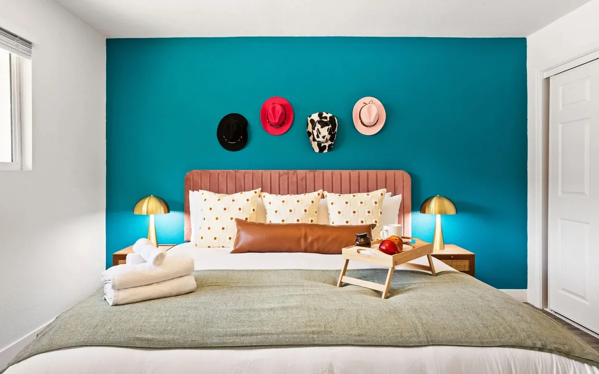A bright bedroom with colorful hats on the wall, a bed with decorative pillows, gold lamps, and a breakfast tray on the bed. The mood is cozy and welcoming, ideal for relaxing or enjoying a quiet morning.