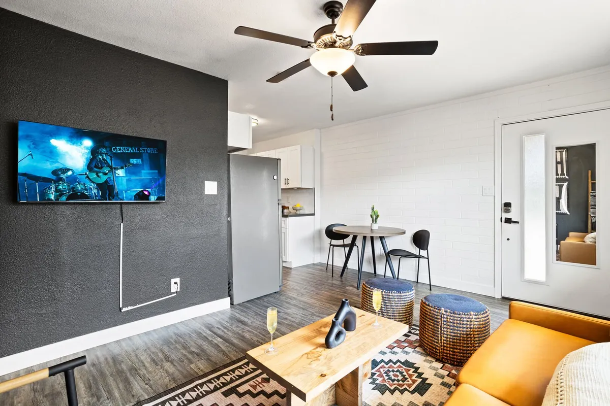 A vibrant living area with an orange sofa, wooden coffee table, TV displaying a band performance, decorative poufs, and a dining area with black chairs. The space is designed for hosting gatherings, watching shows or games, and enjoying drinks or meals together.