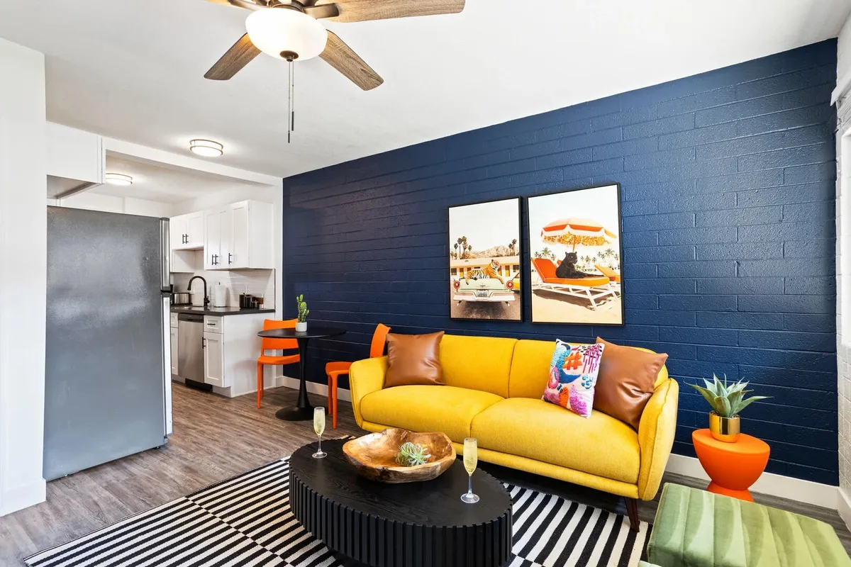 A bright and cheerful living area with a yellow sofa, striped coffee table, and blue accent wall, connected to a modern kitchen area. The room is filled with natural light and offers plenty of seating for group socializing or relaxing.